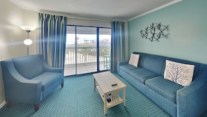 Een kamer met een blauwe bank, een blauwe fauteuil, een klein houten bijzettafeltje en een groot raam met gordijnen.