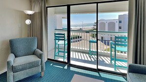 Et hotelværelse med balkon, sofa, stol og udsigt til havet og bygninger.