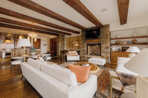 Un salon spacieux avec un grand canapé, une cheminée et un téléviseur. Au fond, on aperçoit une cuisine avec des poutres en bois et un mur en pierre.