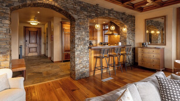 A spacious living area with a stone archway, wooden ceiling beams, a bar area with stools, and a kitchen with stone walls. - Cozy See Forever Condo by InvitedHome | Balcony, Walk to Hike and Festivals (Mountain Village)