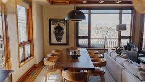 A dining area with a wooden table and chairs, a sofa, and a large window with a view of mountains. - Cozy See Forever Condo by InvitedHome | Balcony, Walk to Hike and Festivals (Mountain Village)