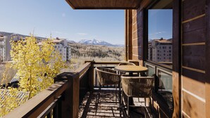 A balcony with a wooden ceiling, wicker chairs, and a small round table. A view of a mountain range and buildings is visible through the window. - Cozy See Forever Condo by InvitedHome | Balcony, Walk to Hike and Festivals (Mountain Village)