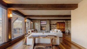 A spacious living room with a fireplace, wooden beams, and a large window. - See Forever Condo by InvitedHome | Ski Valet, Walk to Ski, Hot Tub, Balcony (Mountain Village)