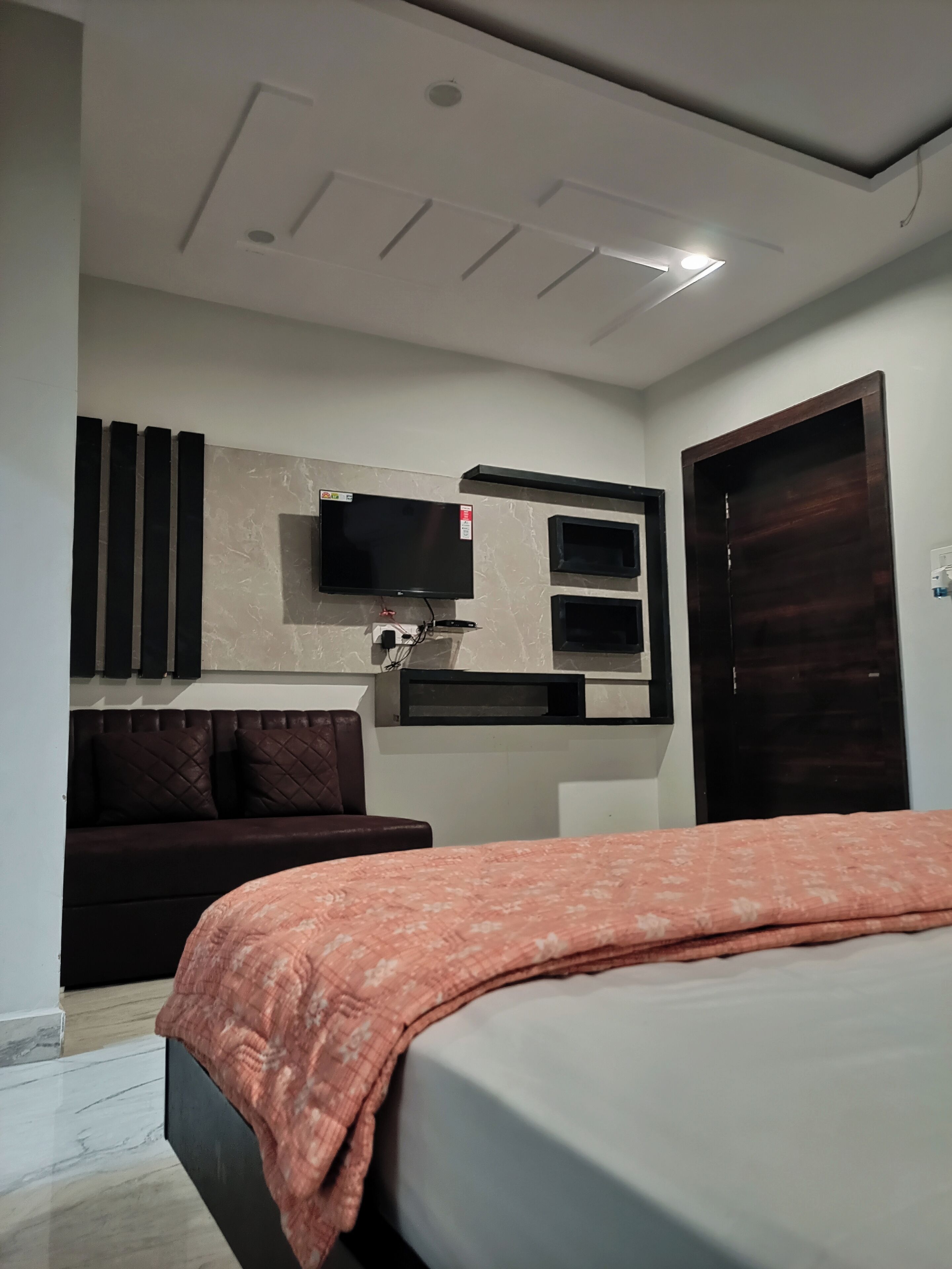Ein modernes Schlafzimmer mit Bett, dunkler Couch, Wand-Fernseher und Holztür.