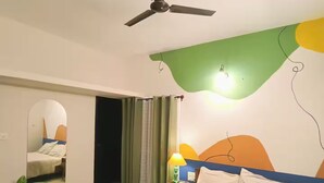 Una habitación de hotel moderna con una cama, un ventilador de techo, un adorno de pared verde, una mesita de noche con lámpara y un espejo.