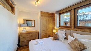 Ein Schlafzimmer mit hölzernem Kopfteil, einem großen Bett mit weißer Bettwäsche, einem Nachttisch mit Lampe, einem Holzschrank und einem Fenster mit Blick auf Gebäude.