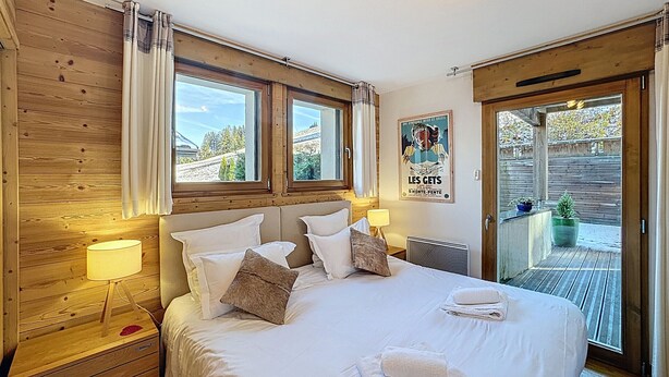 Ein Schlafzimmer mit einem großen Bett, Holzwänden, zwei Fenstern, einem Poster an der Wand und einem Balkon mit Topfpflanzen.