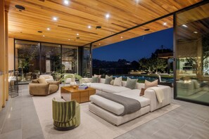 Un salon moderne avec un grand canapé d’angle, une table basse et un fauteuil vert. Un mur de verre offre une vue sur une piscine et des arbres.