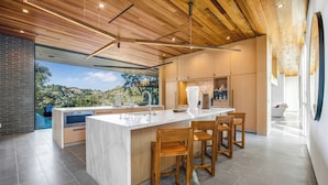 A modern kitchen with a large island, wooden chairs, and a view of a mountainous landscape. - Oak Pass Manor: Indoor-outdoor Luxury With Pool, Sauna & Panoramic Views (Beverly Hills)