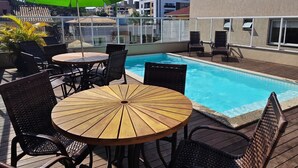 Ein Poolbereich auf dem Dach mit einem Holztisch und Korbstühlen, umgeben von einem grünen Sonnenschirm.