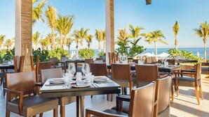 Un área de comedor al aire libre con mesas dispuestas para dos, rodeada de palmeras y con vista a la playa.
