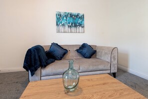 A living room with a beige sofa, two dark blue pillows, a glass vase on a wooden coffee table, and a painting on the wall. - Toast Stays - The Merrington Fold (Kirk Merrington)