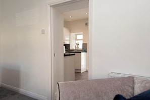 A room with a beige sofa, a white door leading to a kitchen with white cabinets and a window. - Toast Stays - The Merrington Fold (Kirk Merrington)