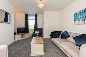 A living room with a beige sofa, a wooden coffee table, a television on a stand, a chair with a cushion, and a door. - Toast Stays - The Merrington Fold (Kirk Merrington)