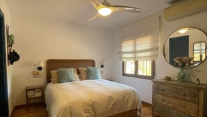 Ein Schlafzimmer mit Bett, Kommode, Deckenventilator, Fenster mit Jalousien und Spiegel an der Wand.