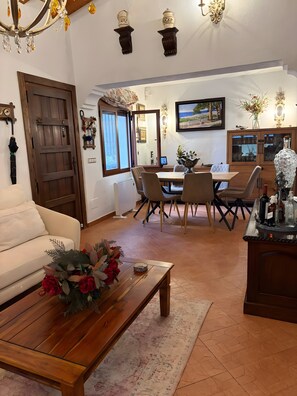 A living room with a sofa, a wooden coffee table with flowers, a dining area with a table and chairs, a kitchen area with cabinets, and a chandelier.