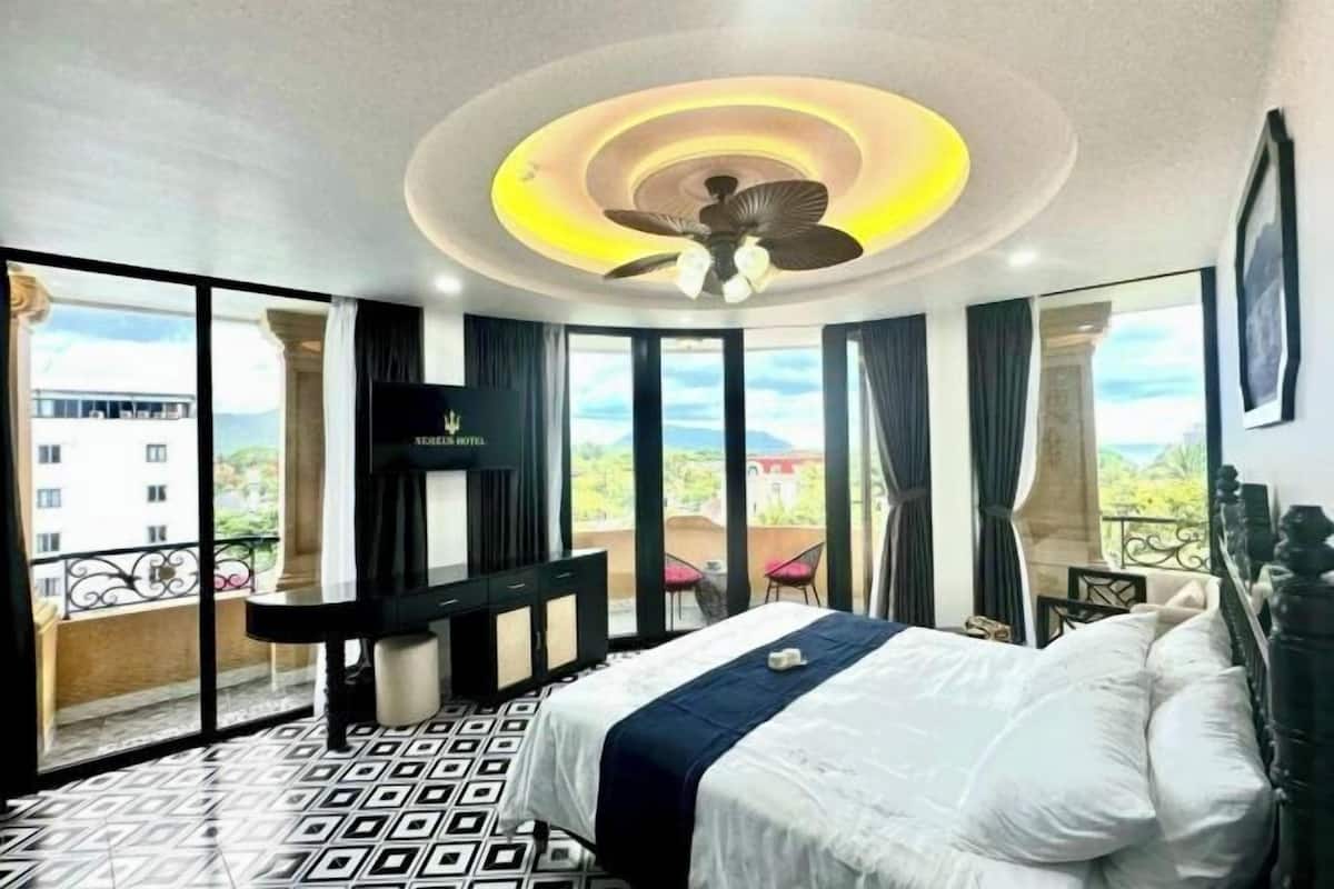 A hotel room with a bed, a ceiling fan, a TV, a balcony with a view, and a patterned floor.