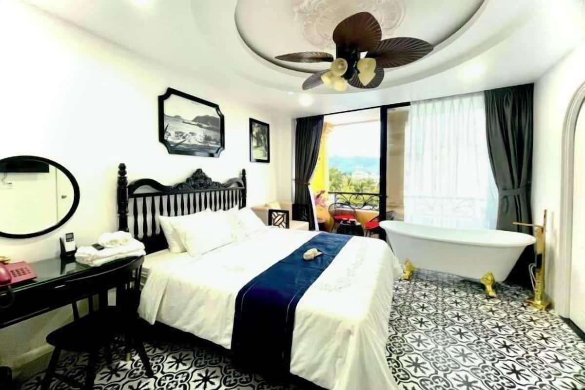 A hotel room with a bed, a round mirror, a telephone, a desk, a bathtub, and a ceiling fan.