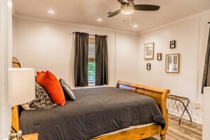 Une chambre à coucher avec un lit, un ventilateur de plafond, une fenêtre avec des rideaux et des tableaux encadrés accrochés au mur.