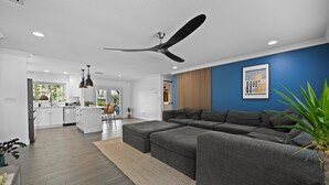 A modern living room with a grey sectional sofa, a ceiling fan, and a kitchen in the background. - The Waterfront Family Oasis- Pool, Dock & Relaxing Backyard (Ellenton)