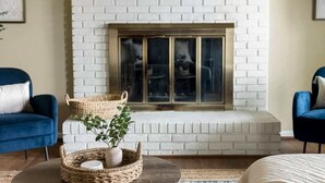 Ein Wohnzimmer mit Kamin aus weißem Backstein, zwei blaue Sessel, einem runden Couchtisch mit Pflanze und einem gewebten Korb.