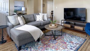 Ein Wohnzimmer mit grauem Sofa, Fernseher, Teppich mit Muster und einem kleinen Beistelltisch.