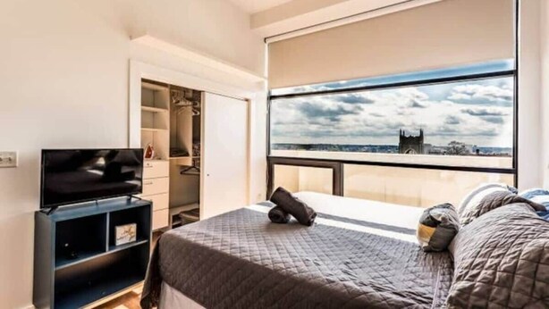 Ein Schlafzimmer mit Bett, Fernseher, Kleiderschrank und einem großen Fenster mit Blick auf die Stadt.