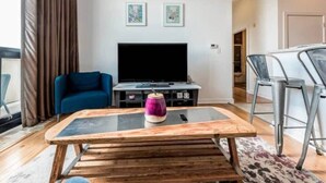 Ein modernes Wohnzimmer mit einem blauen Sofa, einem Glastisch und einem Flachbildfernseher. An der Wand hängen zwei gerahmte Bilder, und eine Tür führt in einen anderen Raum.