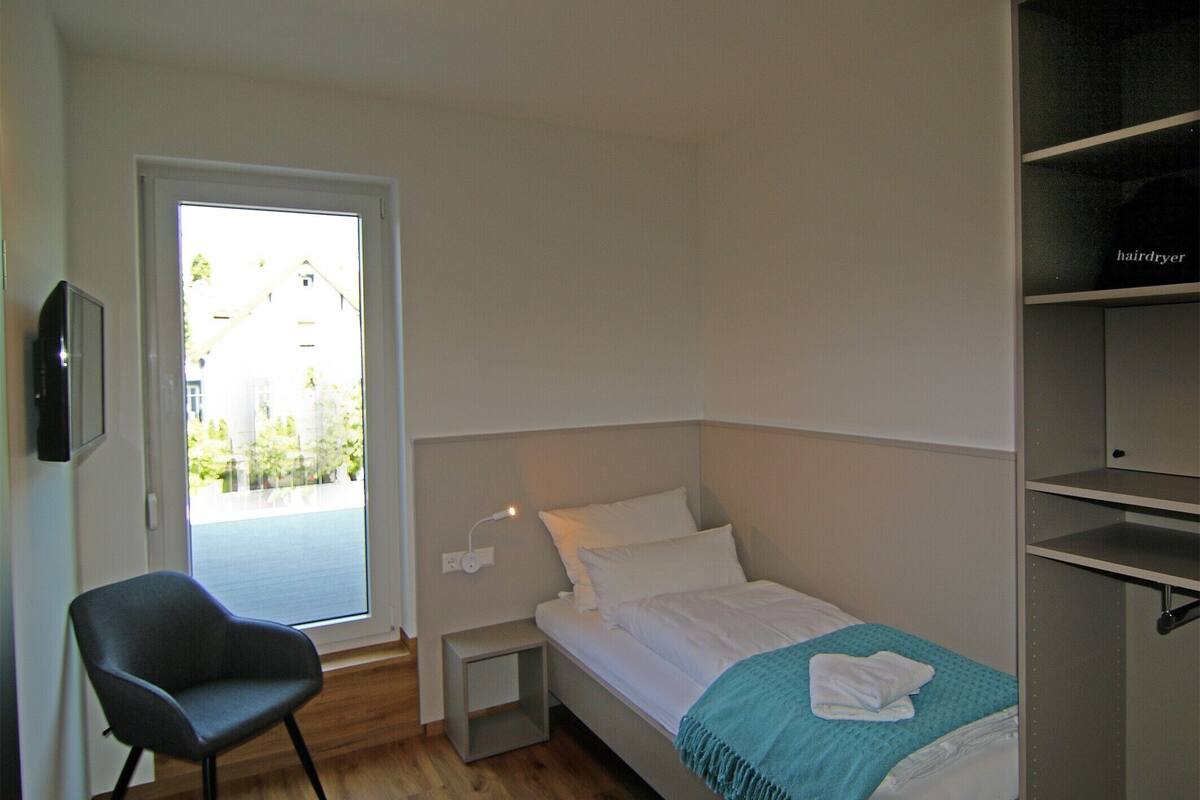 A bedroom with a bed, a chair, a bedside table, and a window.