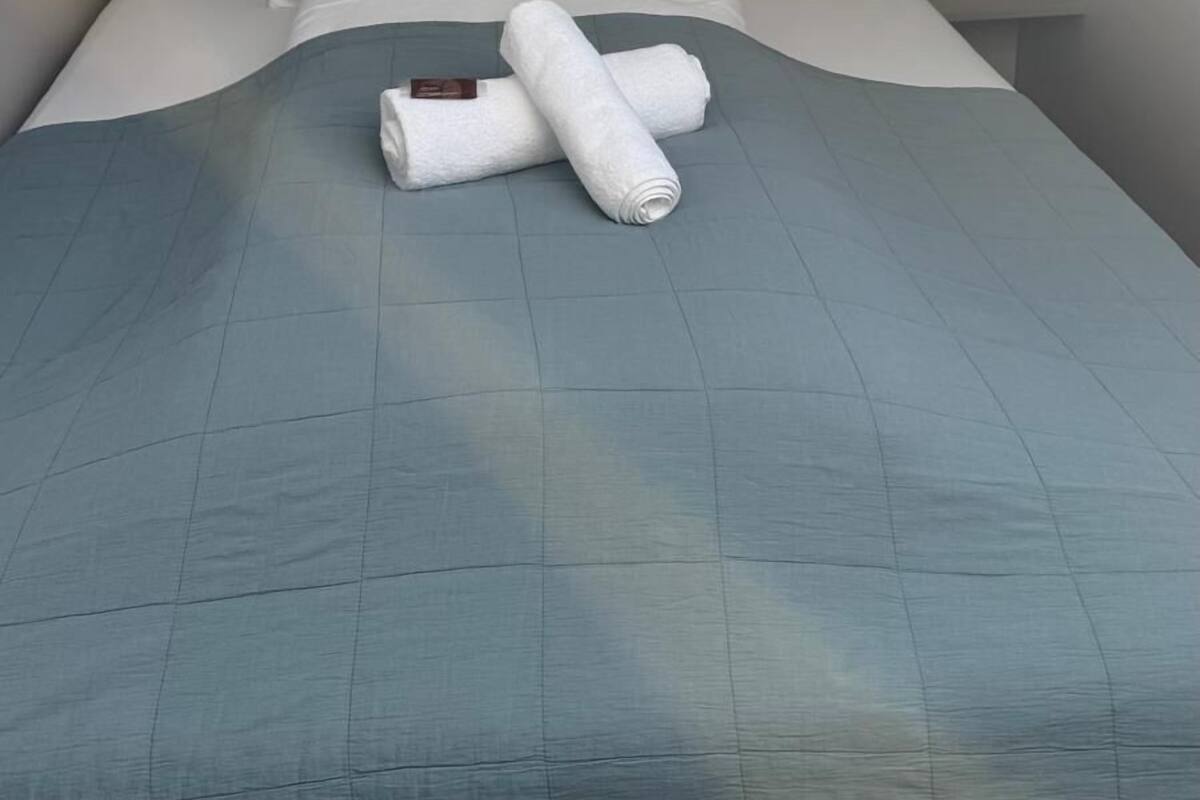 A single bed with a blue quilt, two rolled white towels, and a nightstand with a lamp.