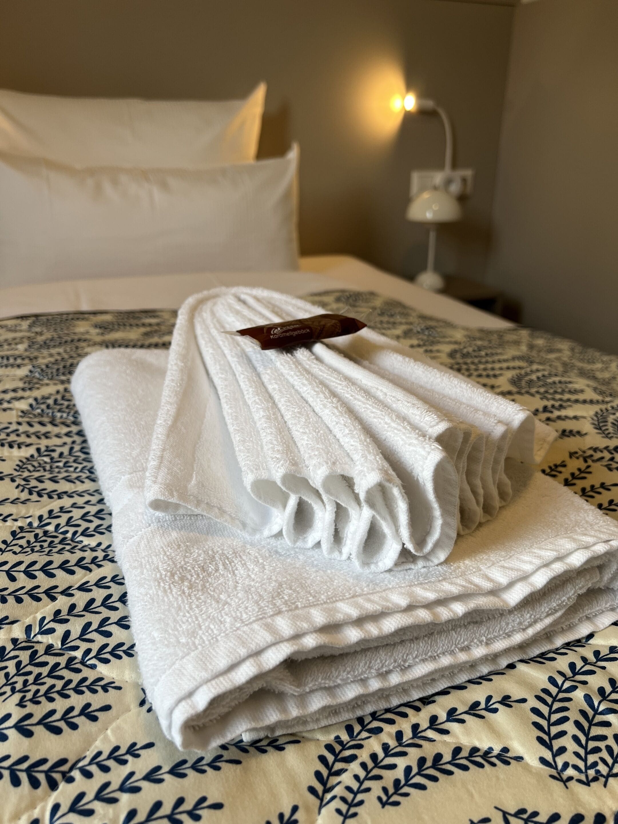 Ein ordentlich gefaltetes weißes Handtuch mit einem Etikett auf einem Bett mit einer gemusterten Bettdecke.