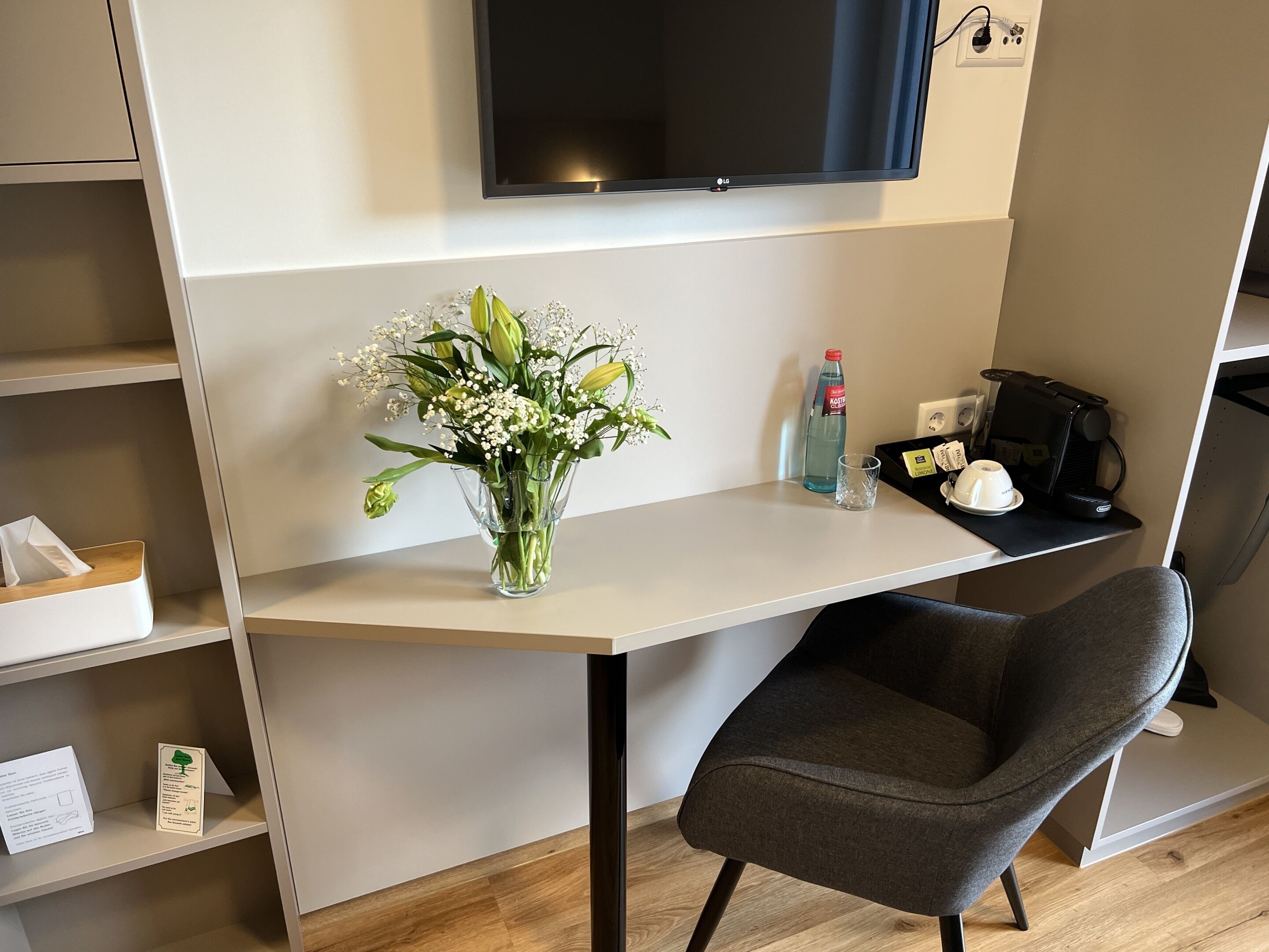 Habitación de hotel moderna con escritorio, una silla, televisor de pantalla plana, un jarrón con flores, una botella de agua, un vaso, cafetera y una bandeja con taza y cuchara.