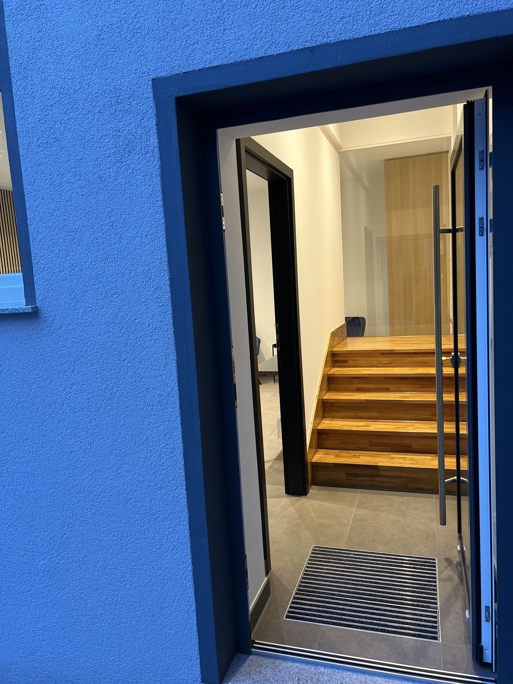 A modern entrance with a glass door leading to a wooden staircase and a small seating area.
