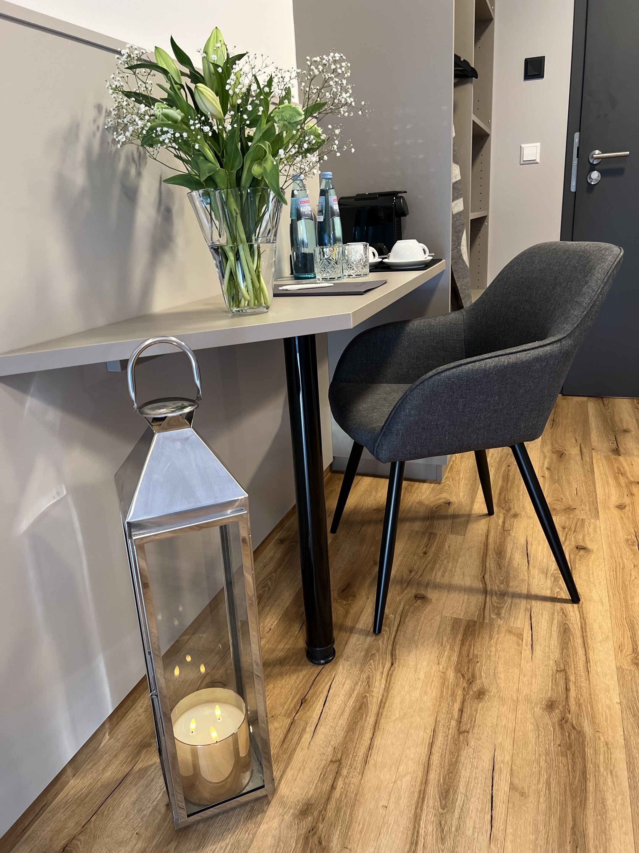 A modern room with a wooden floor, a grey upholstered chair, a small table with a vase of flowers, a candle in a lantern, and a door with a handle.