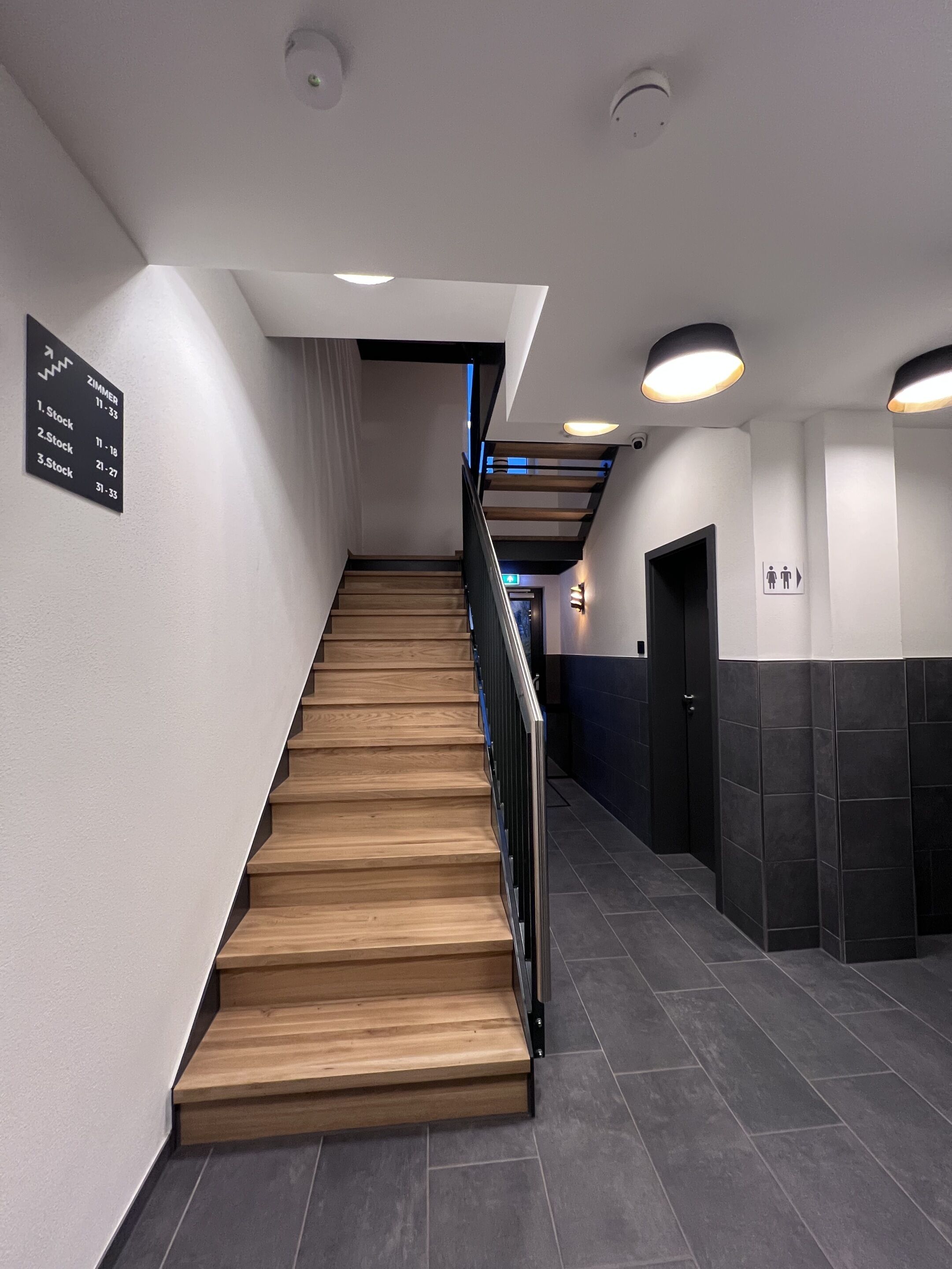 A modern hotel lobby with a wooden staircase, a sign indicating room numbers, and a restroom sign.