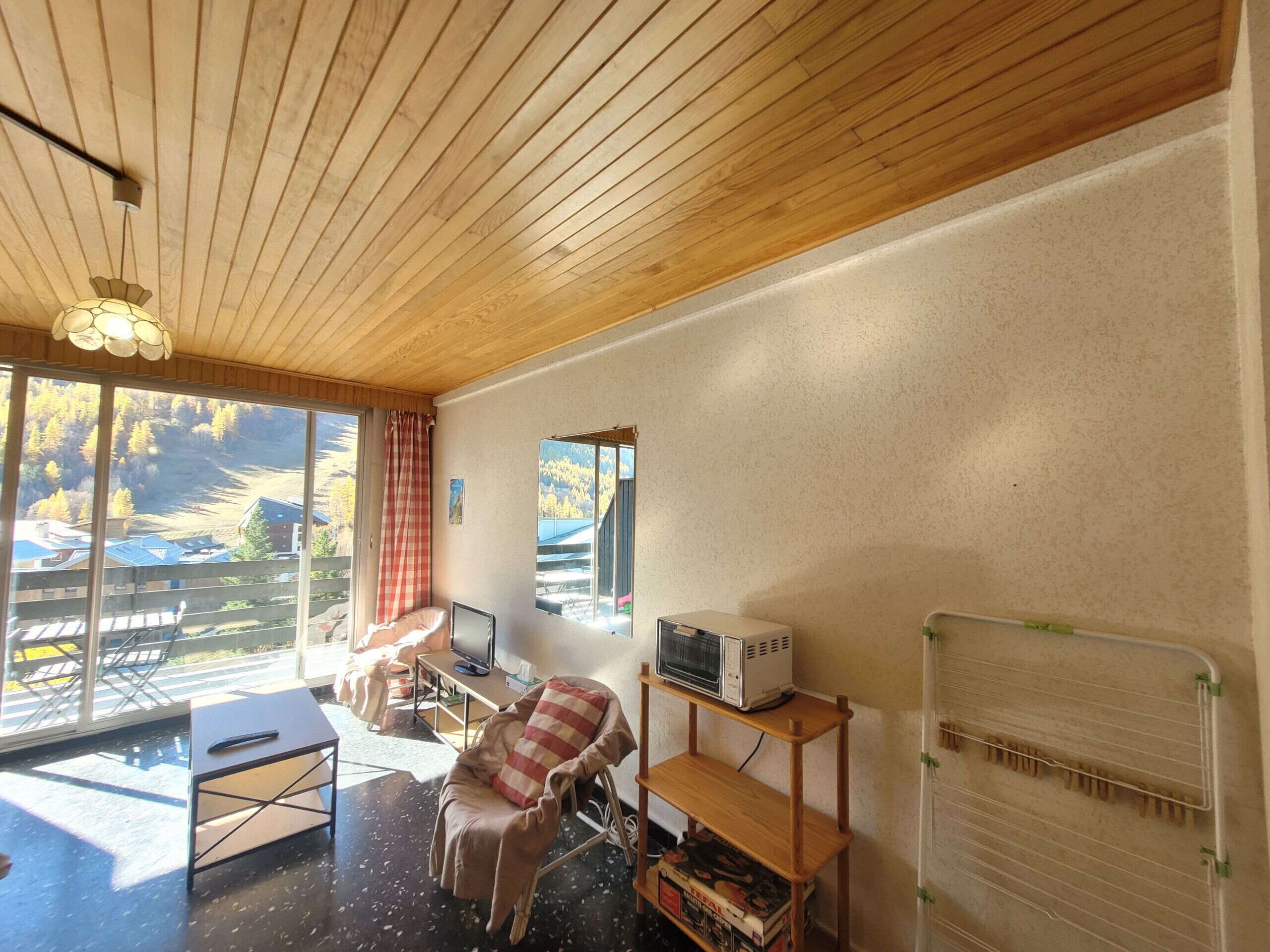Un salon chaleureux avec un plafond en bois, une grande fenêtre donnant sur une forêt, un fauteuil confortable, une petite table et un téléviseur.
