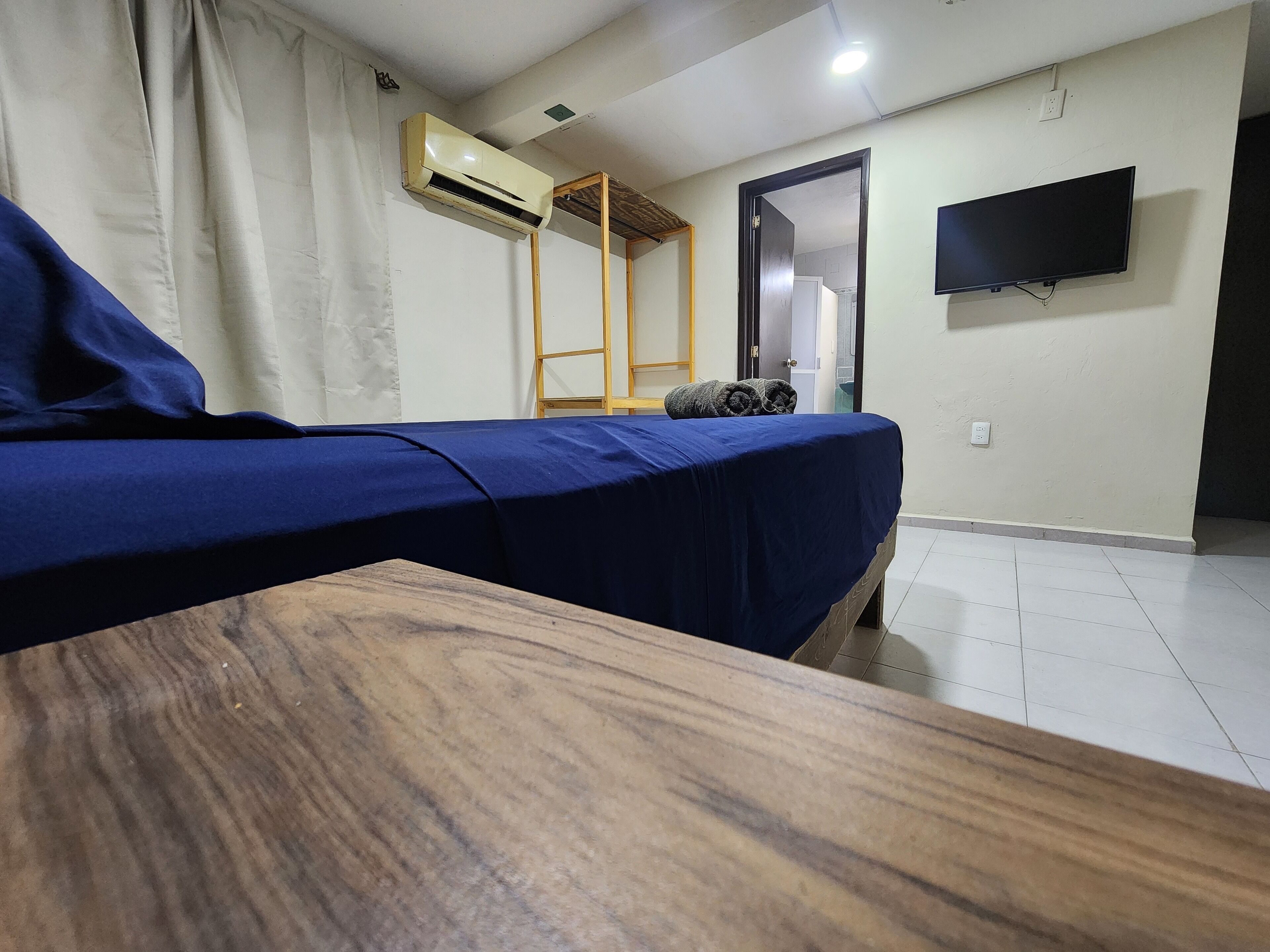 A room with a bed, a wooden table, an air conditioning unit, a TV, and a door leading to another room.
