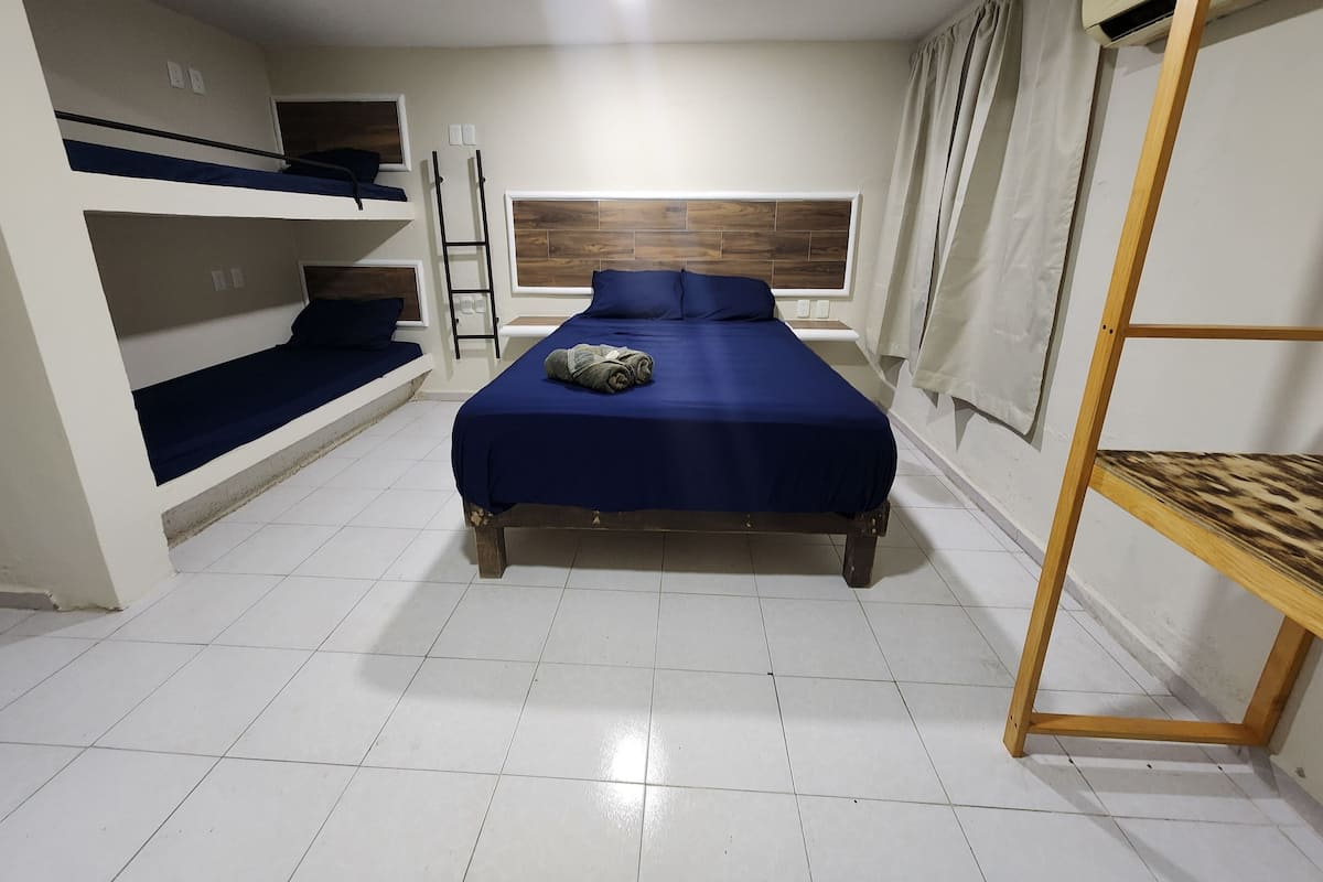 A compact hotel room with a bunk bed, a single bed with a blue bedspread, a wooden shelf, and a window with curtains.