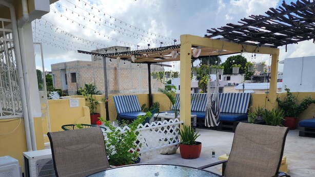 A rooftop patio with a round glass table and chairs, string lights, potted plants, and a pergola. - Casa Seloria (Cancún)