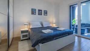 Een moderne slaapkamer met een groot bed, witte nachtkastjes en uitzicht door een glazen deur op een buitengebied.