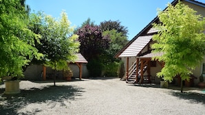 A gravel courtyard with two buildings, one with a staircase and a porch, surrounded by trees and potted plants. - Le Verger du Sausset (Beaune)
