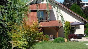 A two-story house with a red roof, a balcony, and a garden with a wooden deck and a small table. - Le Verger du Sausset (Beaune)
