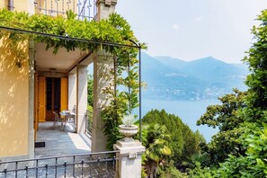 Un balcone con vista su un lago e montagne, un'area pranzo con sedie e tavolo, e una porta che dà su un ambiente interno.