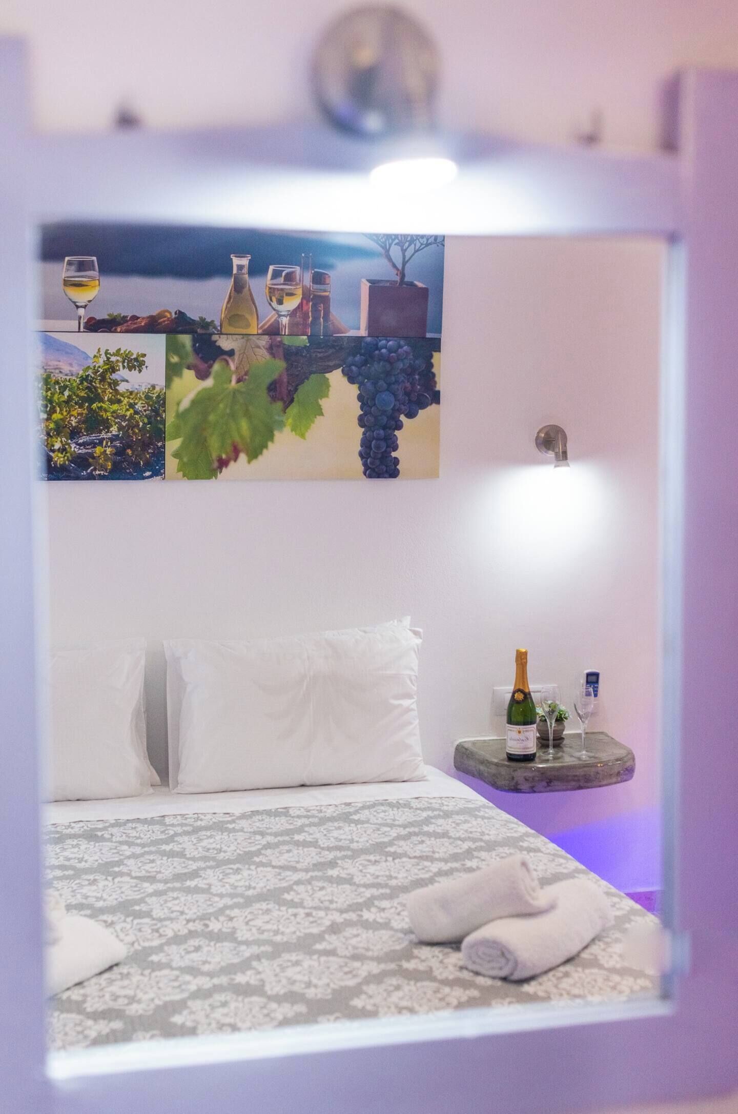 A bedroom with a bed, pillows, a bedside table with a bottle and glasses, and a wall with a painting.