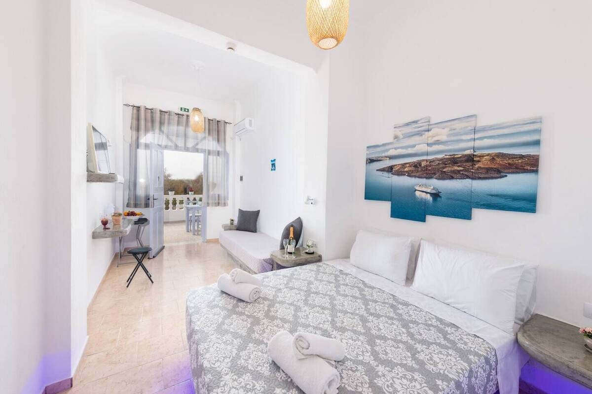 A bedroom with a bed, a nightstand, a window with curtains, a wall-mounted picture, and a balcony with a table and chairs.