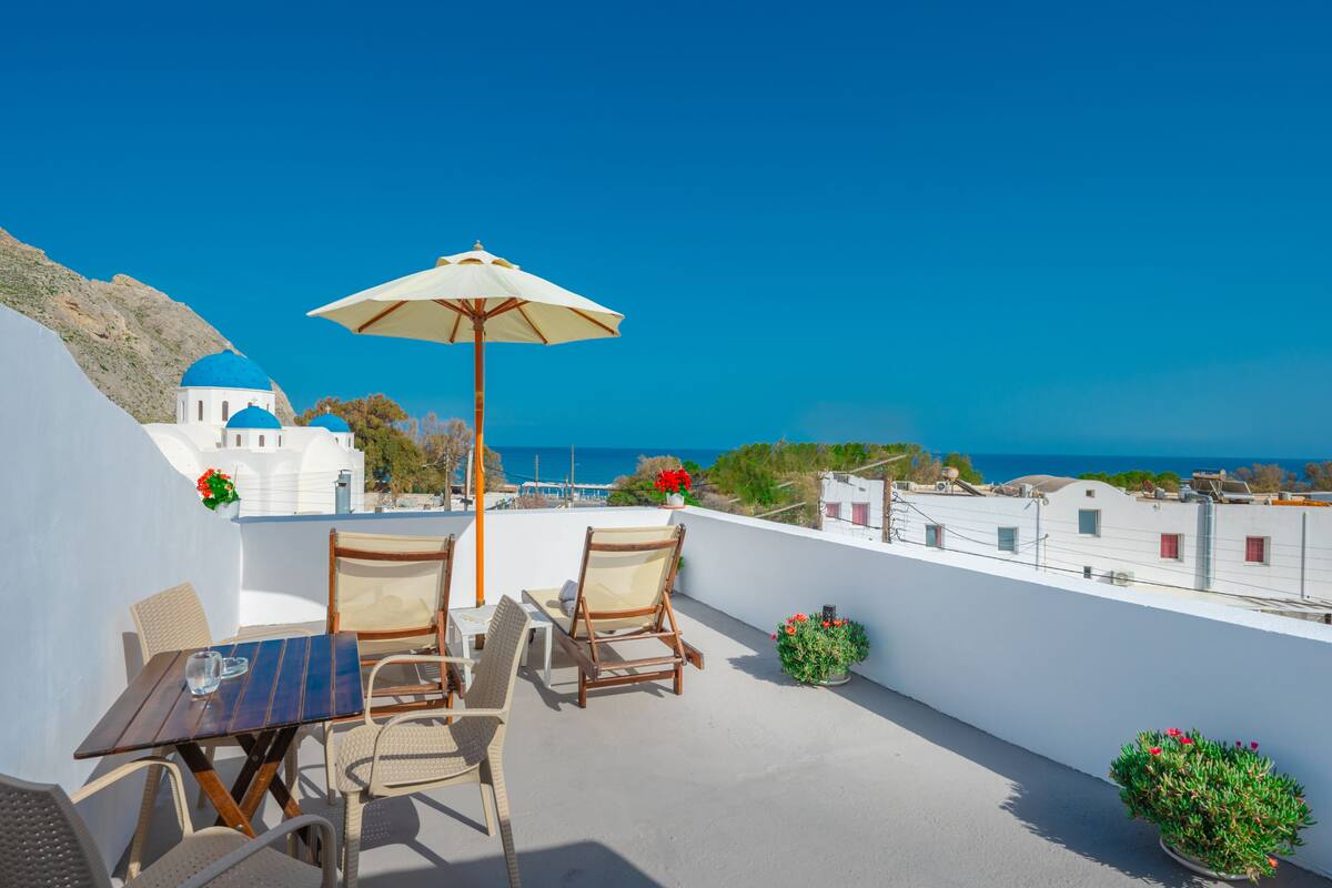 A terrace with a table and chairs, a white umbrella, and a view of the sea and buildings.