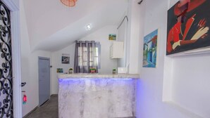 Uma área de recepção com balcão de mármore, porta com grade de metal, janela com cortinas e um quadro na parede.