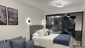 Quarto moderno com cama, sofá cinzento e uma grande janela com vista para a cidade.