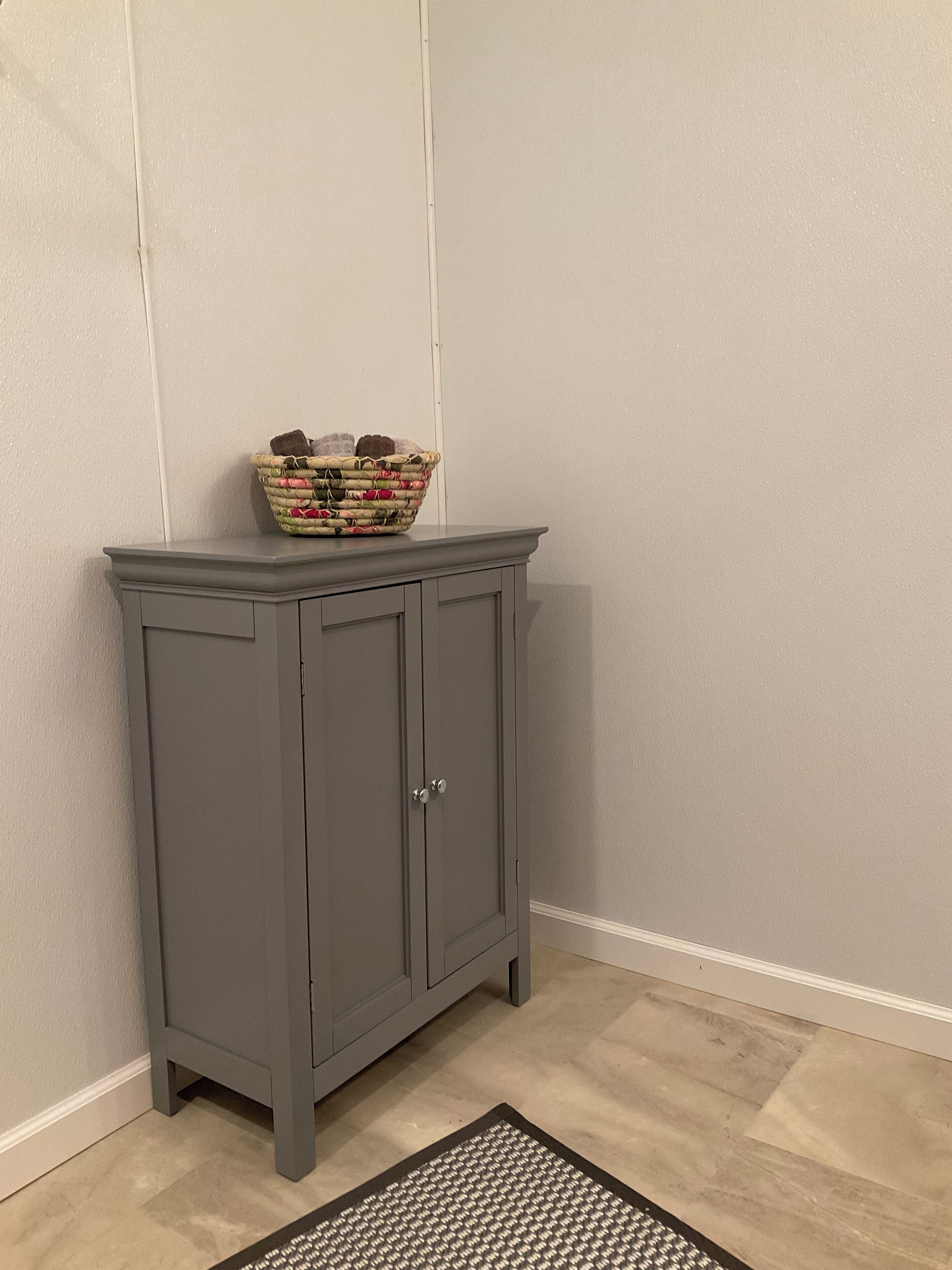 A grey cabinet with a woven basket on top, positioned against a plain wall with a wooden floor.