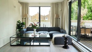 A modern living room with a glass coffee table, a grey sofa, and a view of a balcony with outdoor furniture. - The Westview: 2BR Penthouse &Terrace in White City (London)